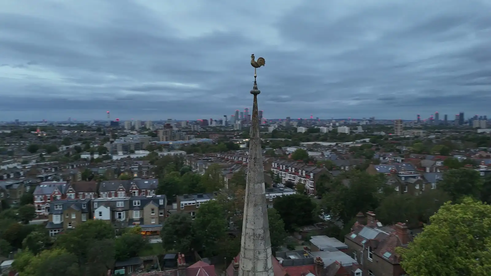 Drone frame level with the church weathercock, sky filling the top half of the frame and the London skyline running along the horizon behind the rooftops.