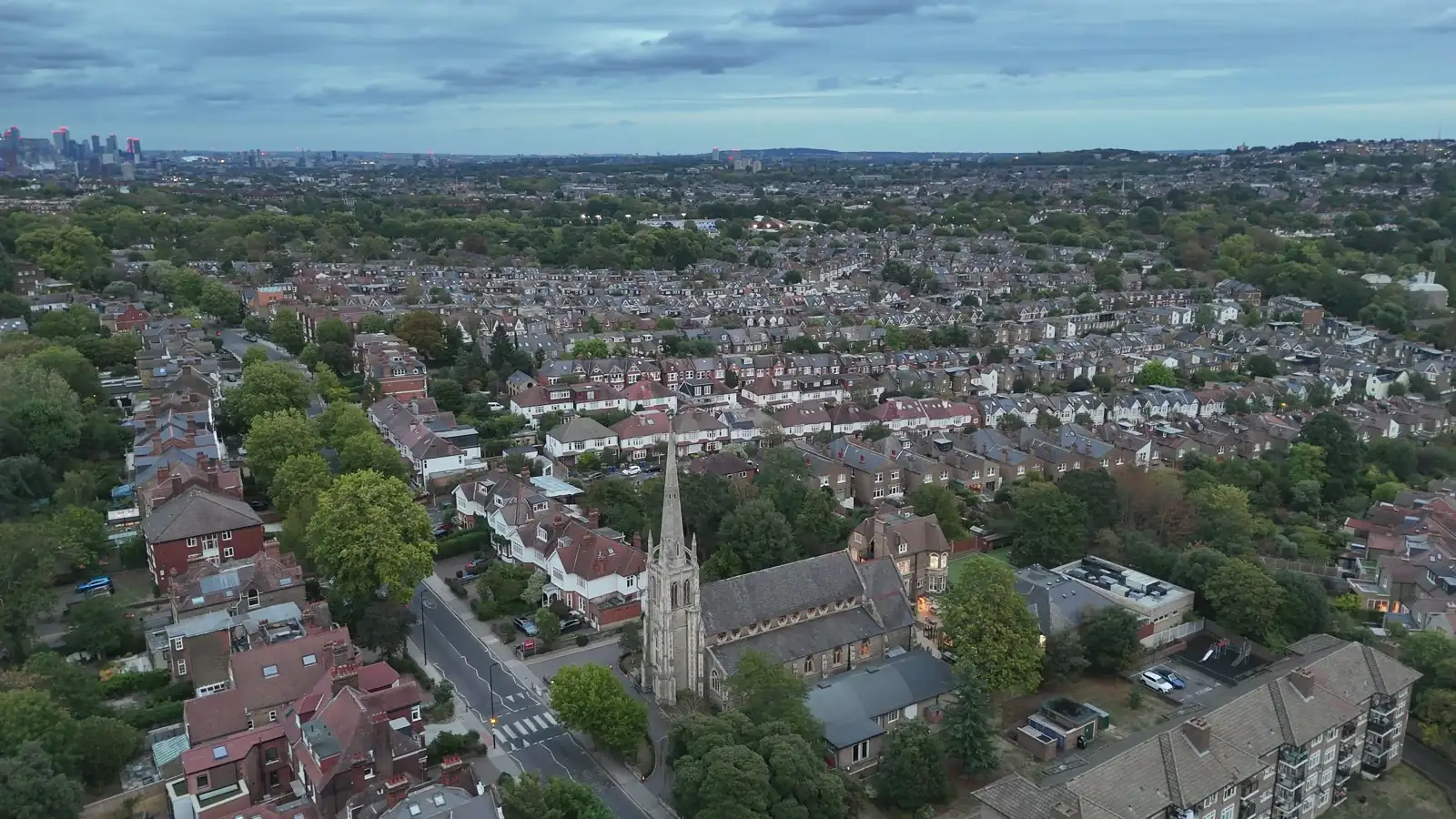 Drone frame high above hundreds of terraced roofs, the London skyline at the top edge, the church and spire occupying about a twentieth of the frame at bottom centre.