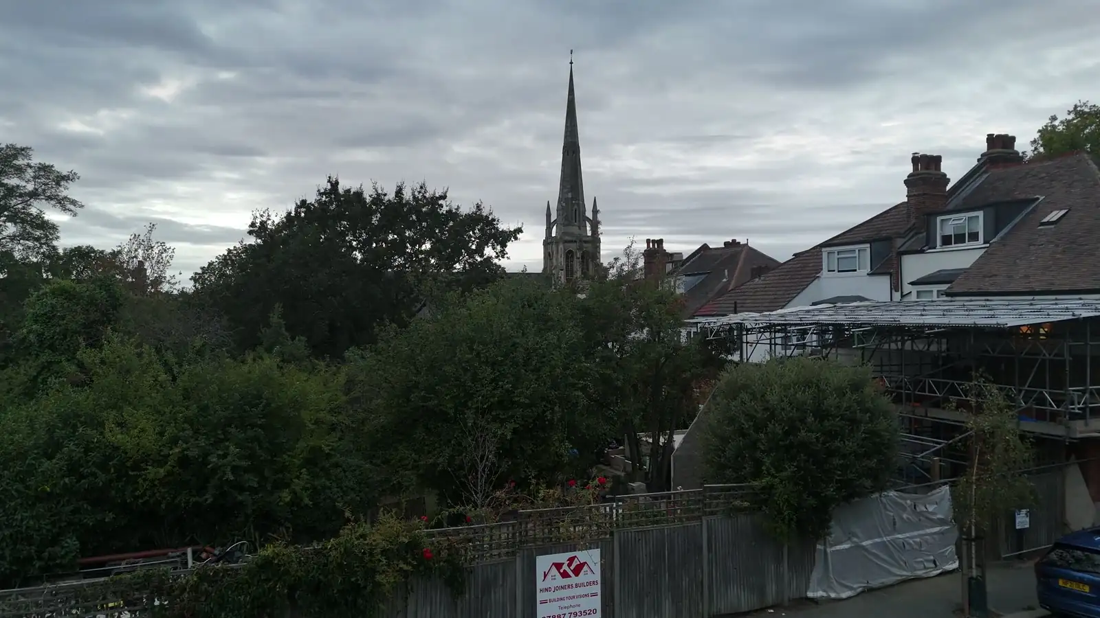 Drone frame shot just after takeoff at about first floor height, looking level across a garden fence and terraced houses, with the church spire small and distant against an overcast sky.