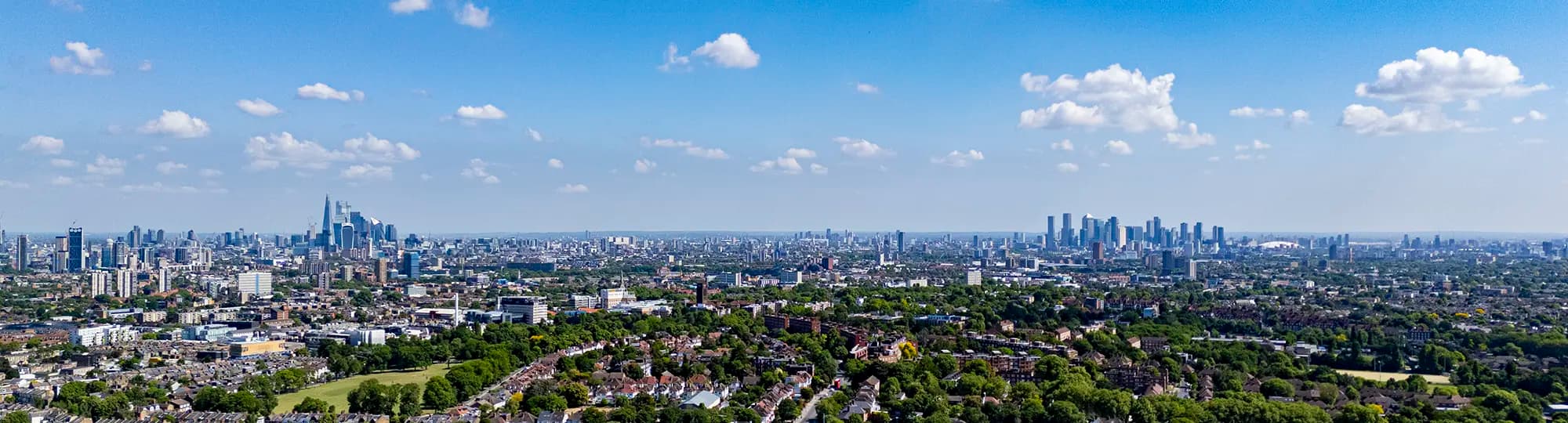 Aerial panorama of London with leafy suburbs and parks in the foreground and the City of London and Canary Wharf skylines on the horizon