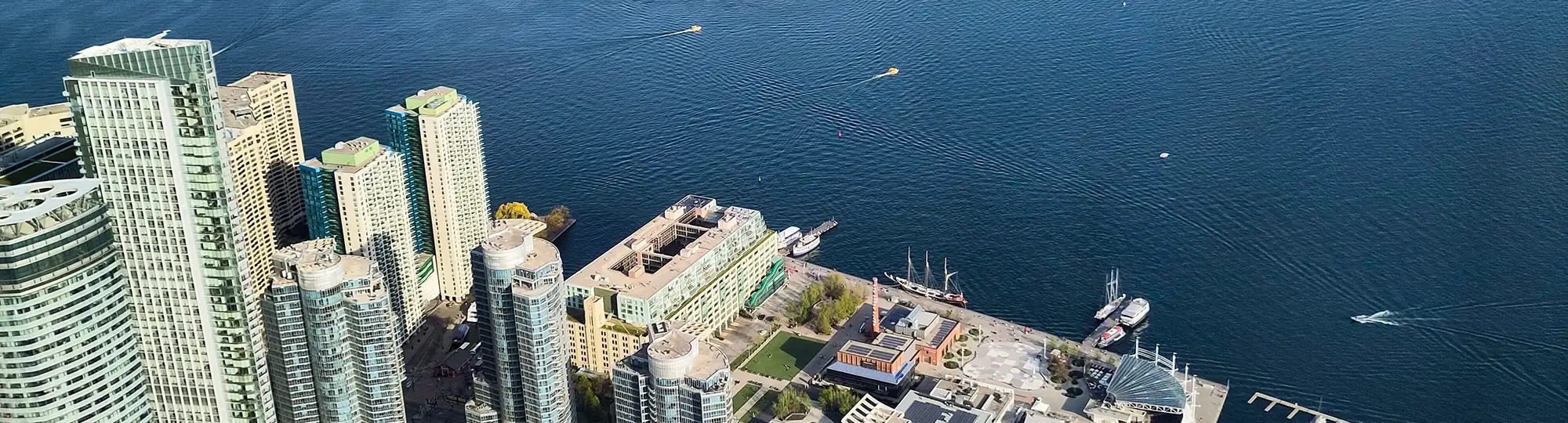 Aerial photograph of the Toronto waterfront looking out over Lake Ontario: a close cluster of tall glass and concrete towers on the left, low quayside buildings, lawns and a tall masted sailing ship moored along the shore, and small boats leaving white wakes across open blue water.